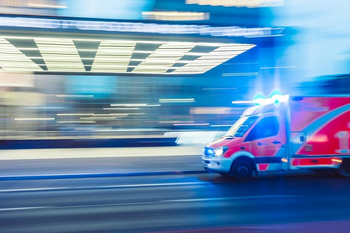 An ambulance with blue emergency lights speeds through a city at dusk