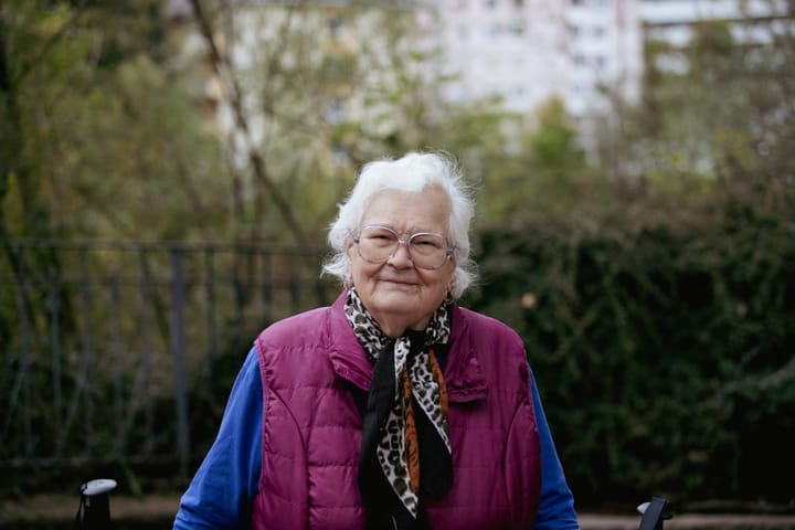 A smiling elderly woman with white hair and glasses sitting outdoors in a park