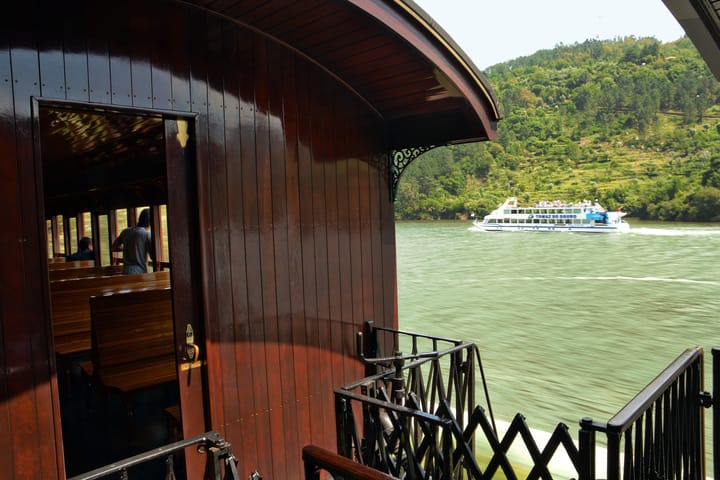 A varnished wooden heritage carriage of the Comboio Historico do Douro beside the Douro river, a tourist cruise boat passing on the water