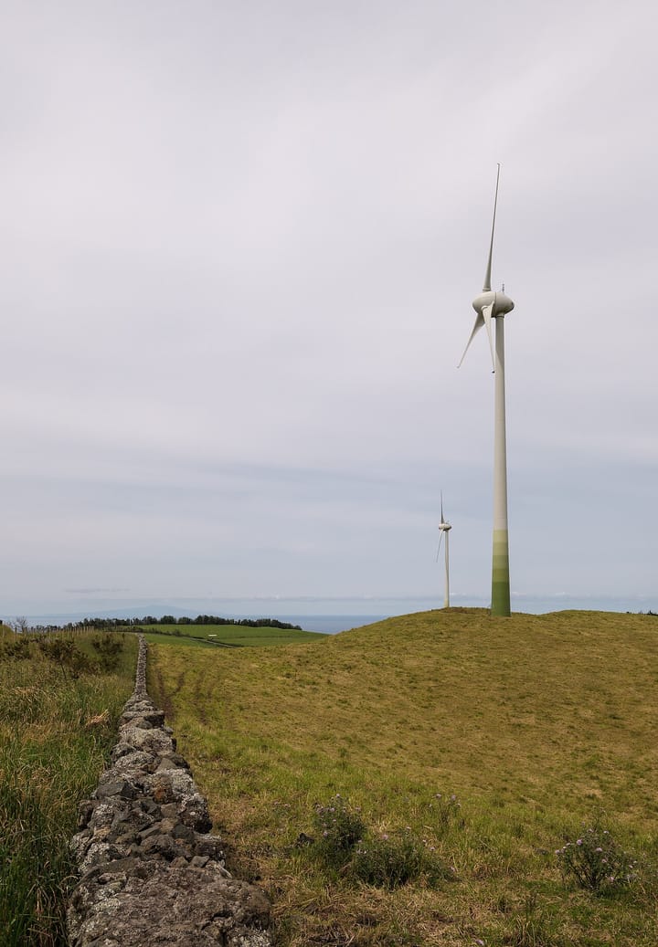 Two wind turbines on a green hillside behind a dry-stone wall at the Serra Branca wind farm in the Azores, Portugal