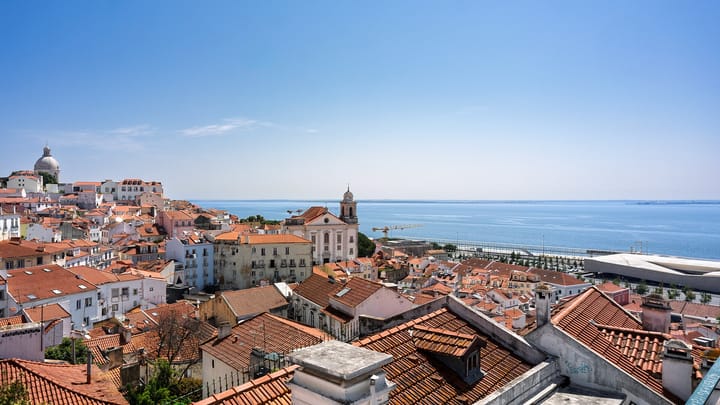 Sunlit terracotta rooftops of Alfama above the Tagus river in Lisbon, Portugal, under a clear summer sky