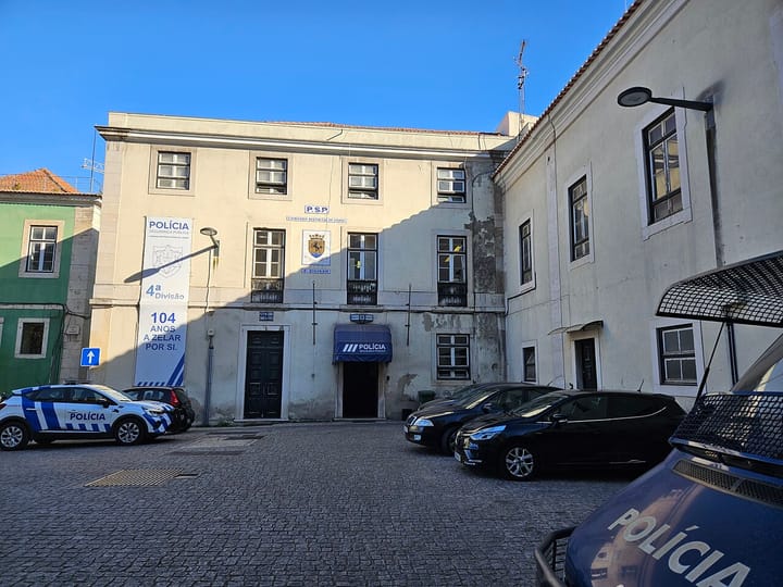 A PSP police station in Alcantara, Lisbon, with blue-and-white marked patrol cars parked on the cobbled forecourt under a clear blue sky
