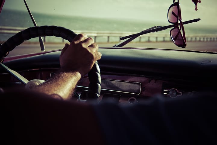 A driver's hand gripping the steering wheel of a car travelling along a coastal road under a hazy sky