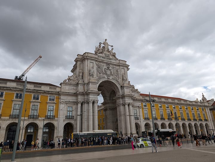 The triumphal Arco da Rua Augusta and the yellow arcaded buildings of Praca do Comercio in downtown Lisbon, with crowds and buses gathered in the square under a cloudy sky