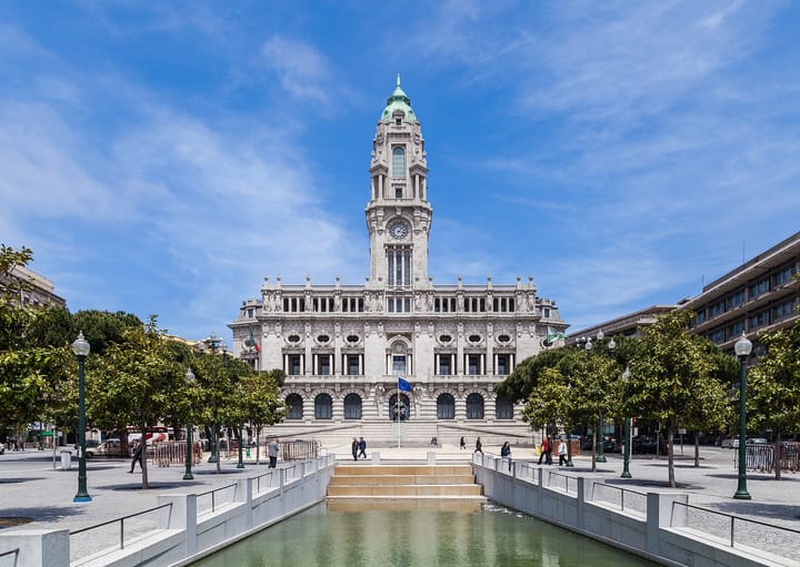 The grey granite facade and green-domed clock tower of Porto City Hall under a blue sky, seen across the reflecting pool of the Avenida dos Aliados