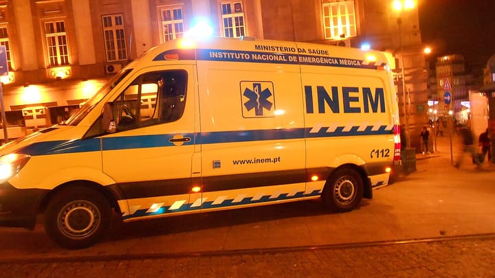 A yellow INEM ambulance with blue lights flashing at night on a Porto street, its side lettering reading Instituto Nacional de Emergencia Medica and 112