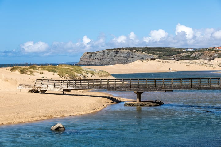 The layered sandstone sea cliff and dune-backed river mouth at Praia da Foz do Sizandro, Torres Vedras, Portugal, with a wooden footbridge crossing the river under a blue sky