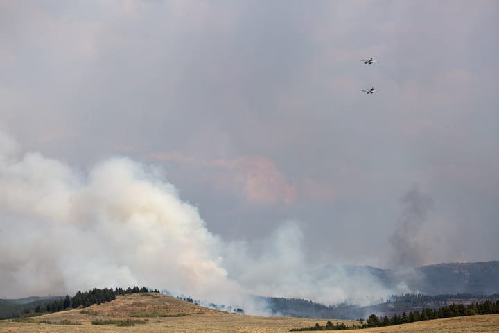 Two water-scooping Fire Boss firefighting aircraft flying in formation over a large plume of smoke rising from a wildfire on forested hills