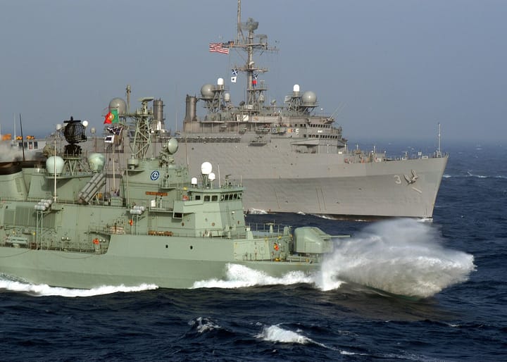 A Portuguese Navy frigate flying the national flag steams at speed through choppy Atlantic seas alongside a larger grey United States Navy warship
