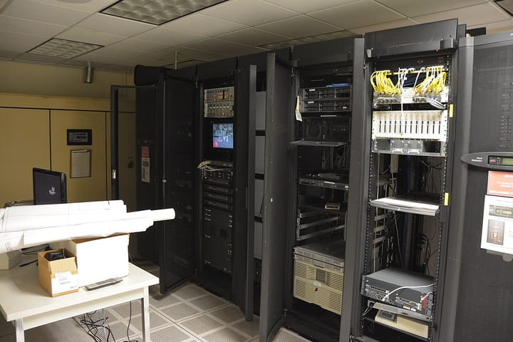 A data-centre server room lined with black equipment racks, network cabling and an APC power-distribution unit