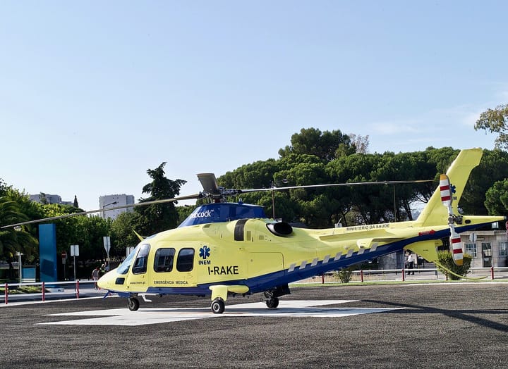 A yellow INEM emergency medical helicopter parked on a hospital helipad in Portugal, with pine trees behind