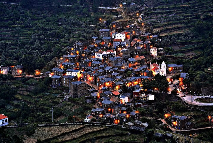 The schist village of Piodao in Portugal's interior at dusk, its dark stone houses clustered on a green terraced hillside with warm street lights glowing among them