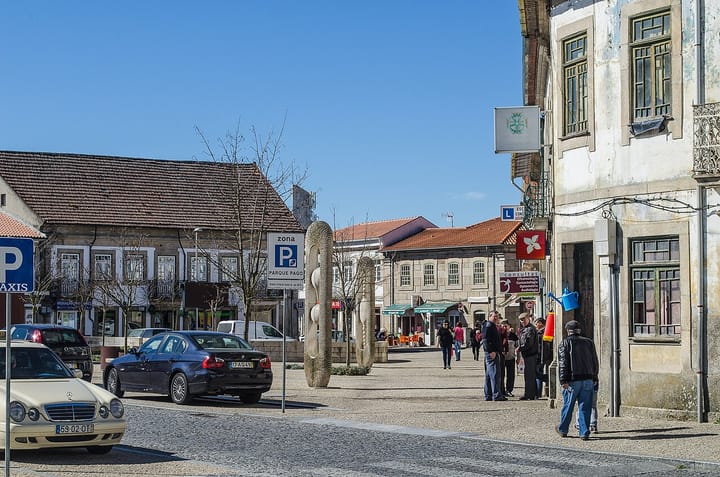 The granite town square of Vila Pouca de Aguiar in Trás-os-Montes, with parked cars, a stone sculpture and residents gathered outside low granite buildings under a blue sky