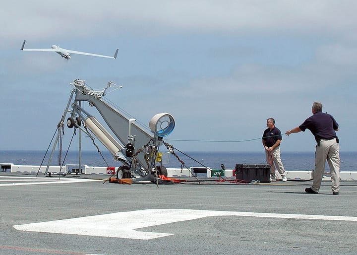 A small fixed-wing surveillance drone launching from a catapult rail as two operators watch, with the open sea in the background