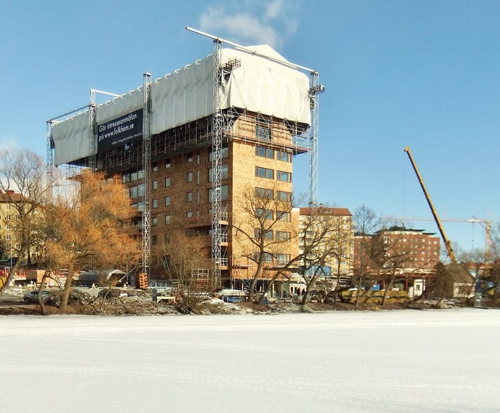A multi-storey timber building under construction, wrapped in scaffolding with a crane alongside, against a clear blue sky