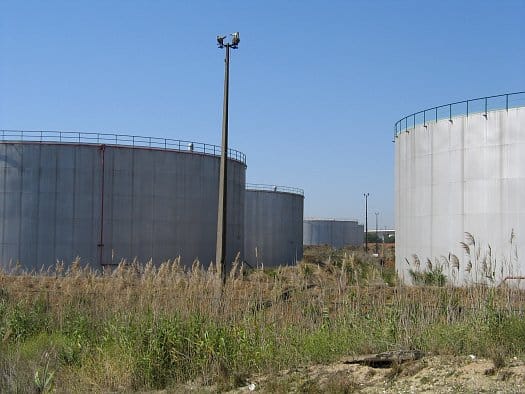 Large cylindrical steel fuel storage tanks at a Galp depot in Portugal, standing behind dry summer grass under a clear blue sky