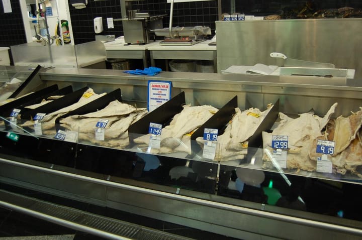 Salted cod (bacalhau) laid out on a chilled counter in a Portuguese supermarket, each pile marked with a printed price and product label
