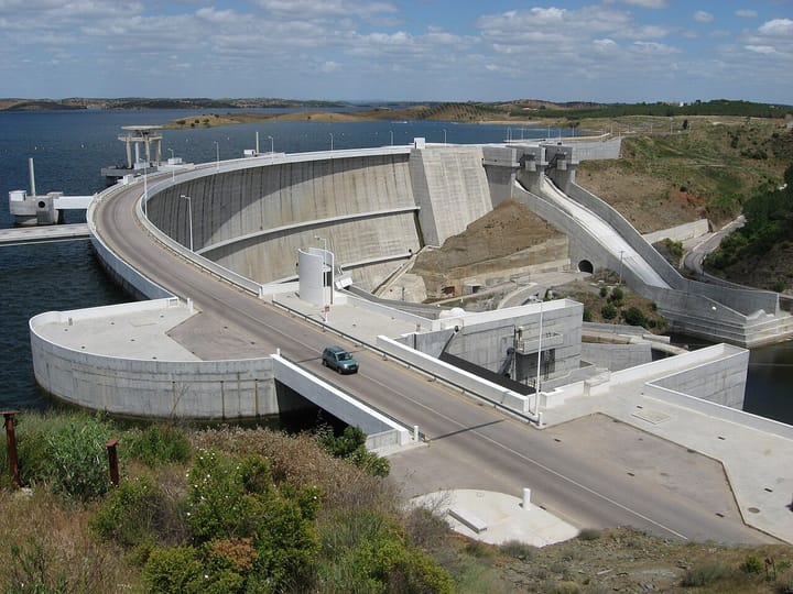 The Alqueva dam on the Guadiana river in Portugal's Alentejo, its curved concrete wall and access road above the reservoir under a blue sky