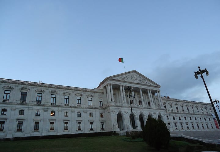 The Palacio de Sao Bento in Lisbon, seat of the Portuguese Parliament, its neoclassical white facade at dusk with the Portuguese flag flying above the central pediment