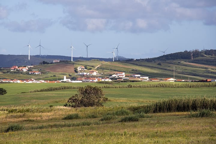 A row of white wind turbines on green hills above a Portuguese village, with farmland in the foreground
