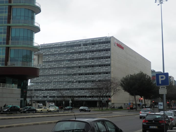 The Vodafone headquarters building at Parque das Nações in Lisbon, its grey scale-patterned façade and red Vodafone logo above a street with parked cars