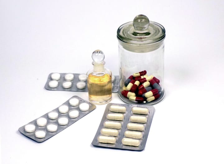 Blister packs of white pills and red-and-cream capsules beside a glass apothecary jar and a small bottle of medicine on a white background