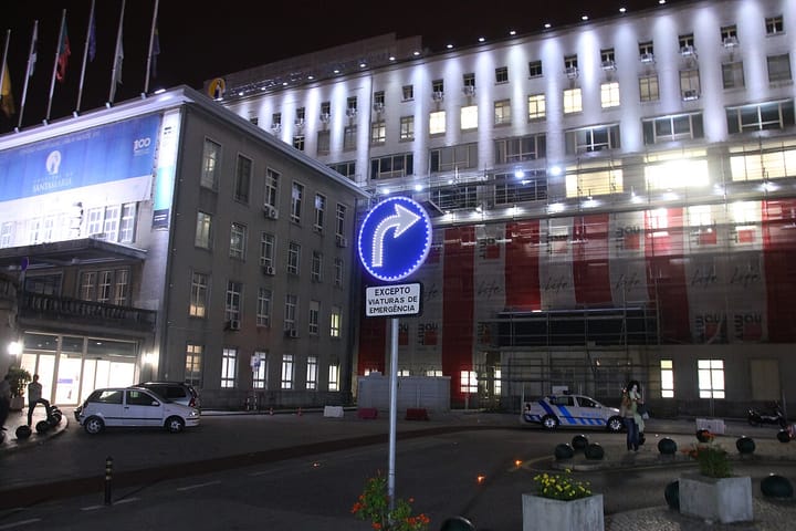 The Hospital de Santa Maria in Lisbon lit up at night, with an illuminated Santa Maria banner, a PSP police car and an emergency-vehicles-only road sign in the forecourt