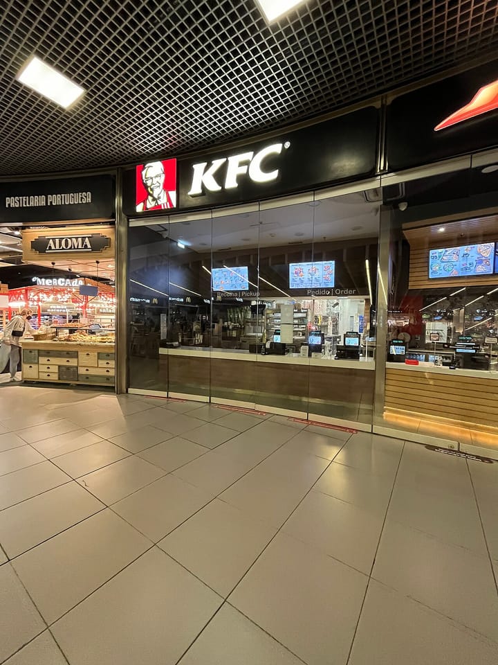 A KFC quick-service restaurant unit inside a Portuguese shopping concourse, its illuminated sign and glass front beside a Portuguese pastry counter