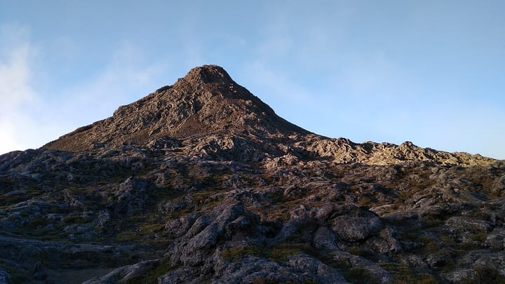 The bare volcanic summit cone of Pico mountain in the Azores, Portugal's highest peak, rising above a rocky lava landscape under a clear evening sky