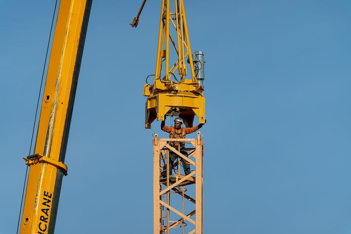 A construction worker in a hard hat and safety harness atop a tower crane against a clear blue sky