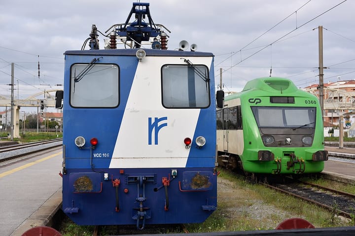 Two Comboios de Portugal trains, a green passenger railcar and a blue maintenance locomotive, standing side by side at a station platform