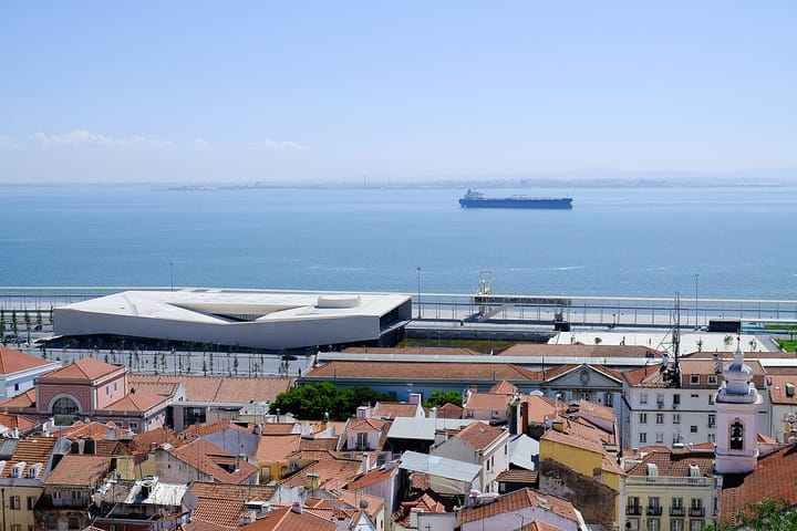 View over the terracotta rooftops of Lisbon's Alfama towards the white curved Lisbon Cruise Terminal on the Tagus riverfront, with a ship on the water