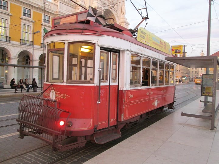 A classic red Lisbon tram parked at Praca do Comercio at dusk, with yellow buildings and tram tracks curving away in the background