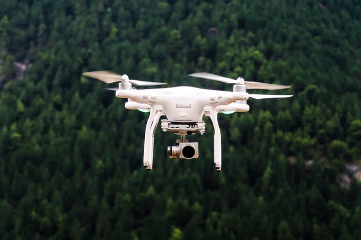 A white quadcopter drone with a camera gimbal hovering in flight in front of a dense green pine forest
