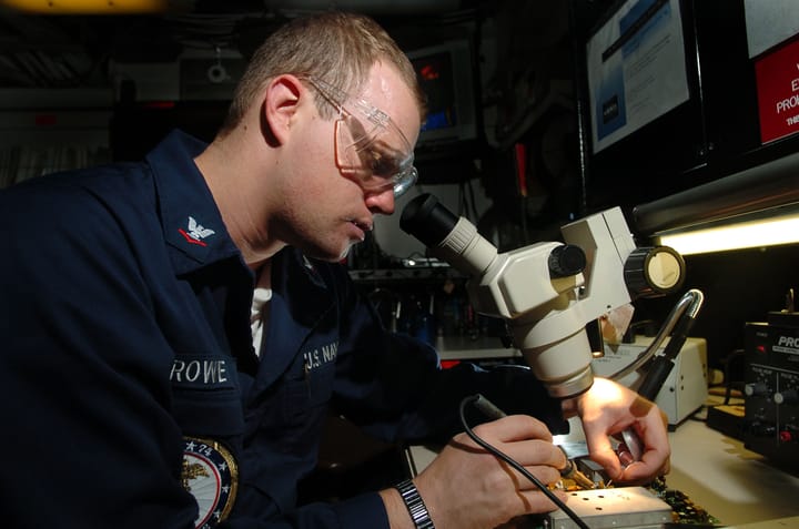 A technician in safety goggles uses a soldering iron and microscope to repair a green electronic circuit board at a workbench