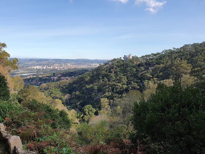 A lush green forested valley of the Serra de Sintra, with the Pena Palace silhouette on a wooded hilltop and the town and coastal plain visible below under a blue sky