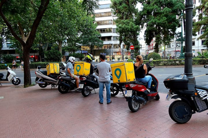 Glovo food-delivery couriers with yellow branded boxes parked on scooters beside a city street