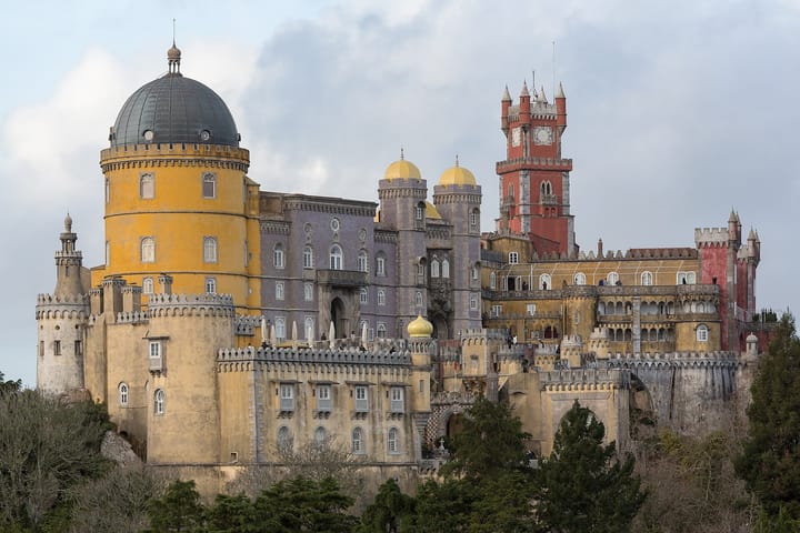 The colourful yellow and red towers of the Pena Palace rising above wooded hills in Sintra, Portugal, under a pale sky