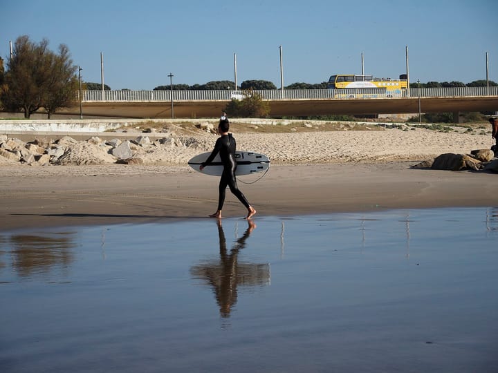 A surfer in a black wetsuit carries a surfboard across the wet sand of Matosinhos beach in northern Portugal, with a road bridge behind