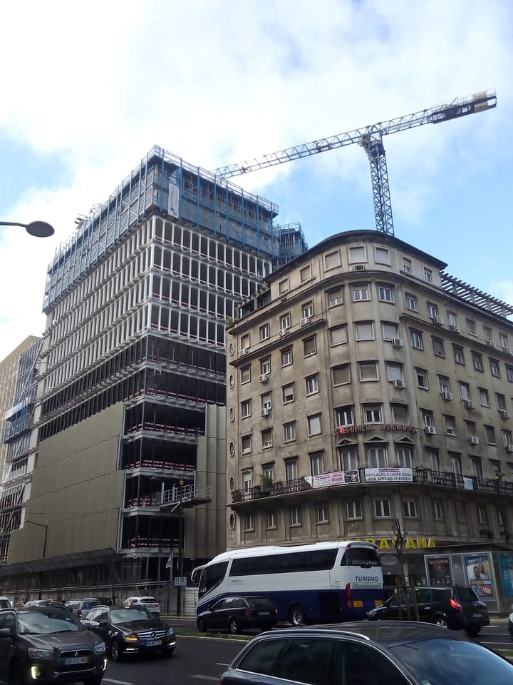 A multi-storey building under construction behind a tall crane, next to an older apartment block, on a street in Lisbon, Portugal