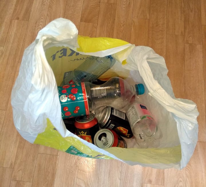 A translucent plastic supermarket bag filled with empty clear PET drink bottles and used aluminium cans on a wooden floor, ready to be returned for recycling