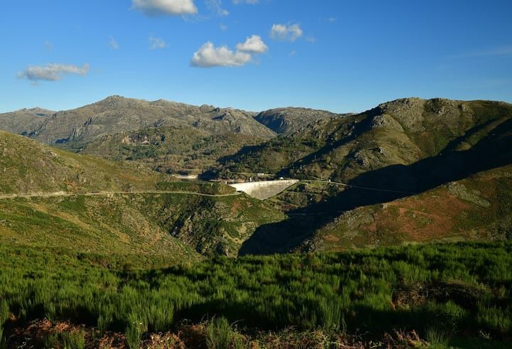 The concrete arch of the Alto Lindoso dam and its reservoir seen from a hillside across a green granite valley in northern Portugal, under a blue sky.