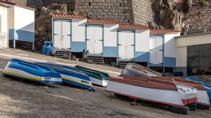 Small wooden fishing boats hauled up and turned over on the slipway at Camara de Lobos, Madeira, in front of a row of white and blue fishermen's huts.