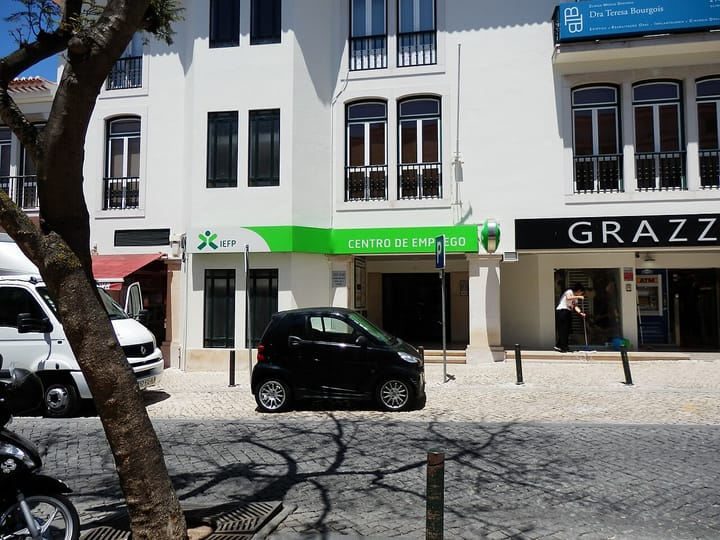A white Portuguese building in Cascais with a green IEFP Centro de Emprego sign above the entrance, a small black car parked at the kerb and cobbled pavement in the foreground