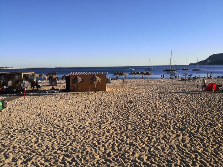 Praia de Albarquel in Setúbal, one of the five contested beaches, with thatched sun umbrellas, bathers, yachts moored in the Sado estuary and the Arrábida hills beyond.