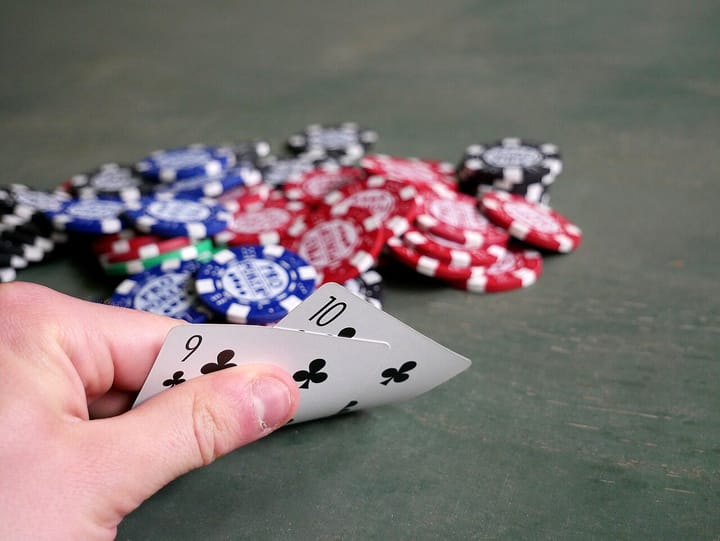 A hand holding two playing cards beside a pile of red, blue and black poker chips on a green table, representing gambling