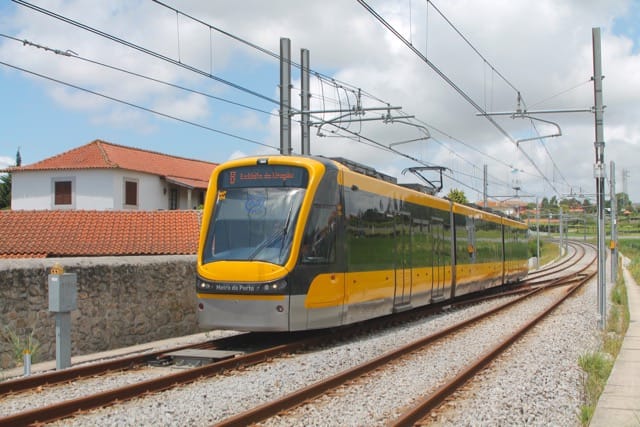 A yellow and grey Metro do Porto tram running on an open-air track under a bright summer sky, bound for Estadio do Dragao