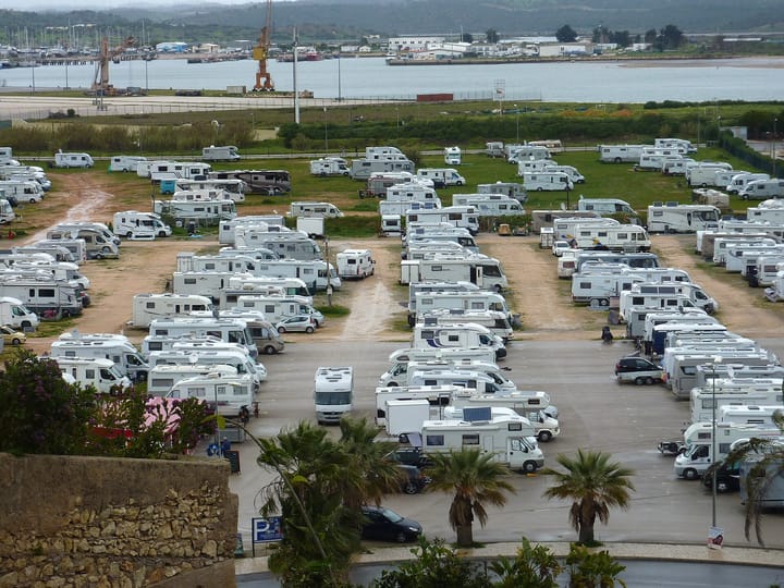 Rows of white motorhomes parked on gravel and grass at a motorhome area beside the marina at Portimao in the Algarve, with the river and port cranes behind