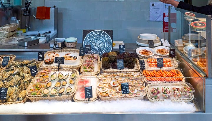 A seafood counter inside Porto's Mercado do Bolhão, with oysters, mussels, scallops, prawns and sea urchins laid out on ice behind handwritten price cards