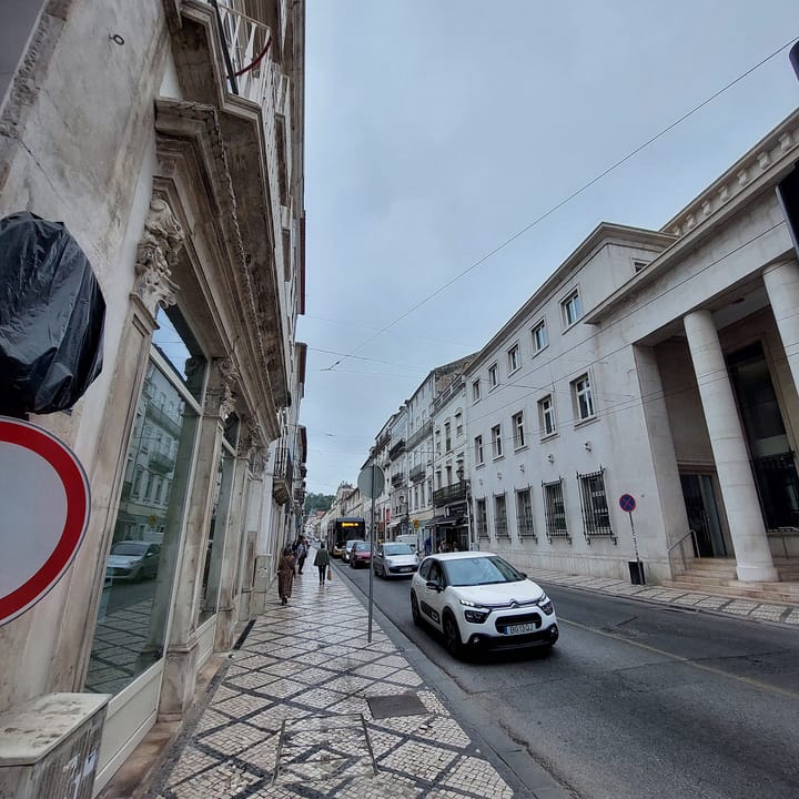 An SMTUC bus and parked cars on Rua da Sofia in Coimbra's Baixa, under trolleybus wires, with the colonnaded Igreja de Santa Justa building on the right